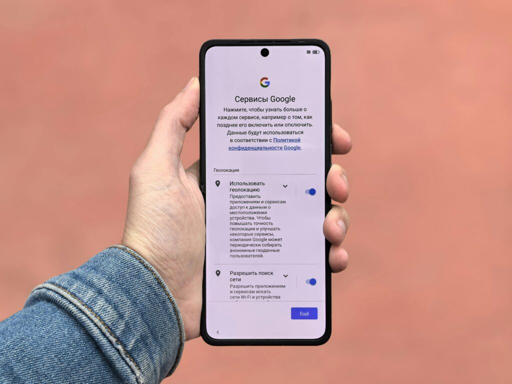 A person holding a smartphone displaying Google settings on a simple background.